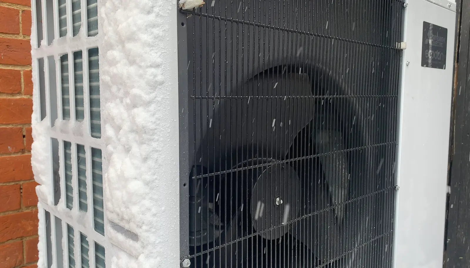 Grille du ventilateur d’une unité extérieure de pompe à chaleur, au pied d’un mur, par temps de neige